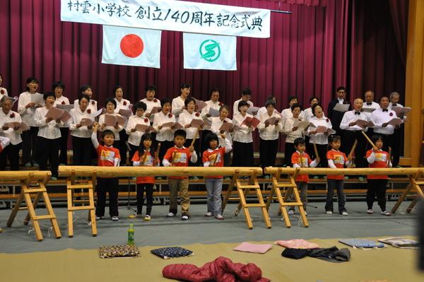 村雲小学校 創立140周年記念式典と書かれている横断幕のある舞台の上で、白いシャツに胸に赤い花をつけている合唱団の方が歌を歌っており、舞台下には、オレンジ色のティーシャツを着た子どもがバチを持ち太鼓のように設置された柱を叩いている様子の写真
